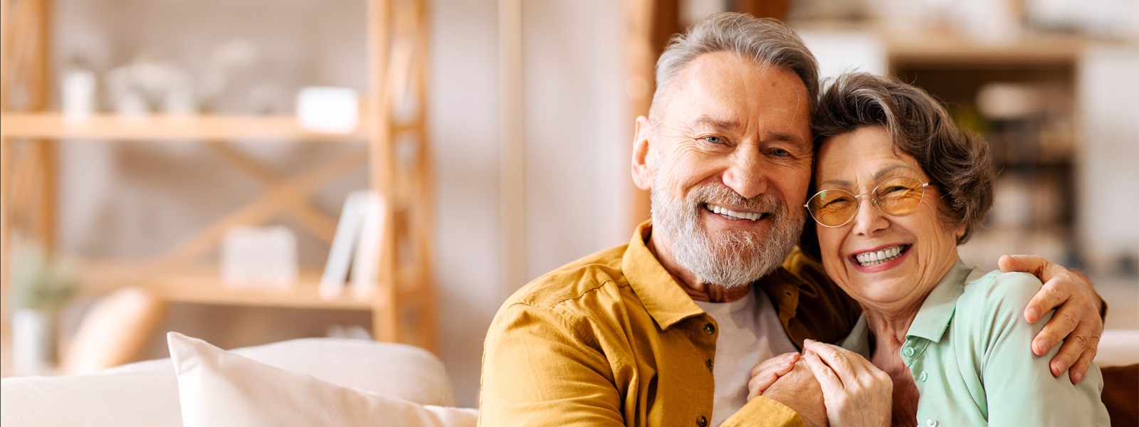 Happy elderly partners. Senior man and woman sharing tender moment, hugging and radiating happiness, sitting on sofa in living room and smiling at camera